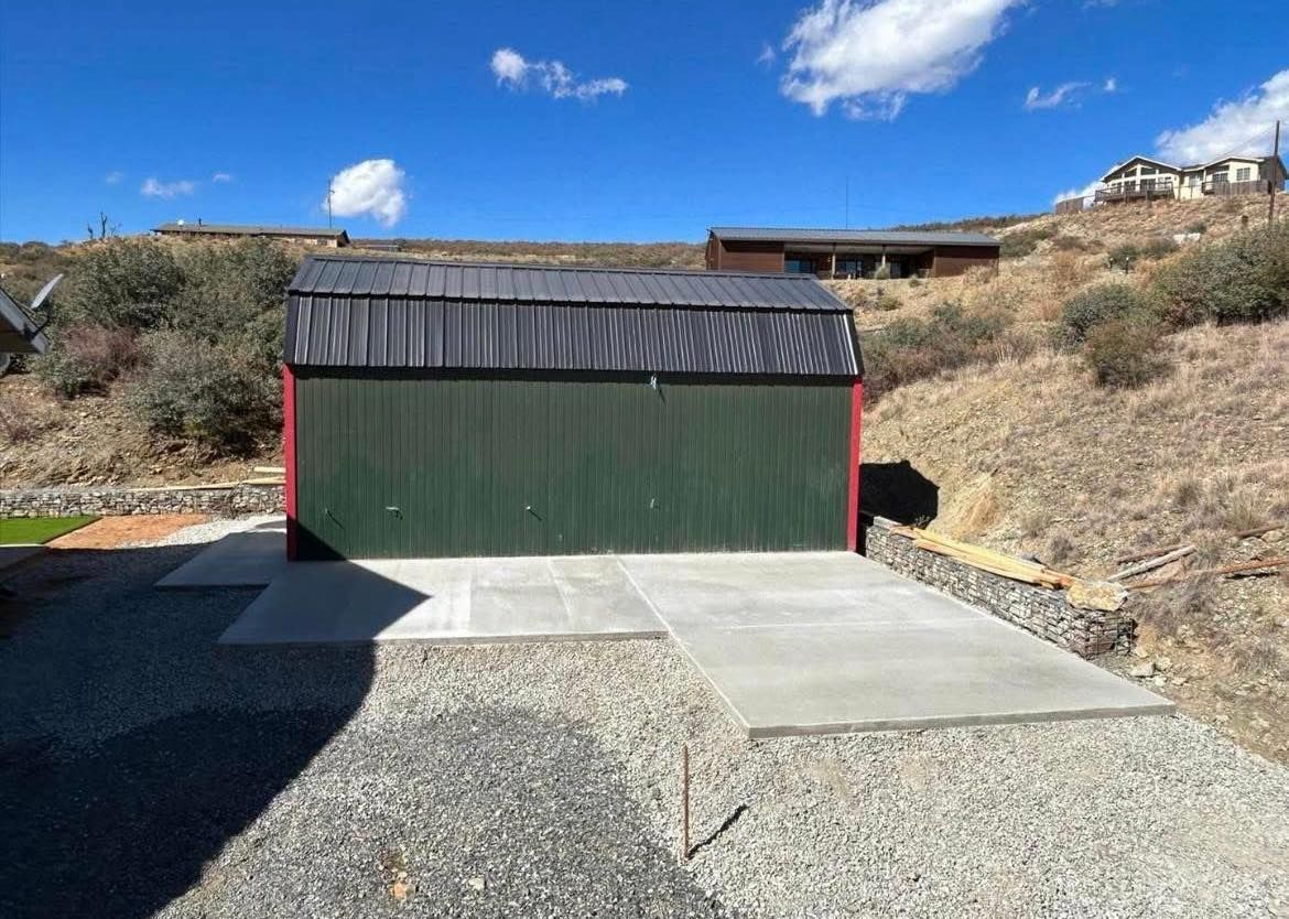 Green corrugated metal shed with concrete driveway on a hillside under a blue sky.