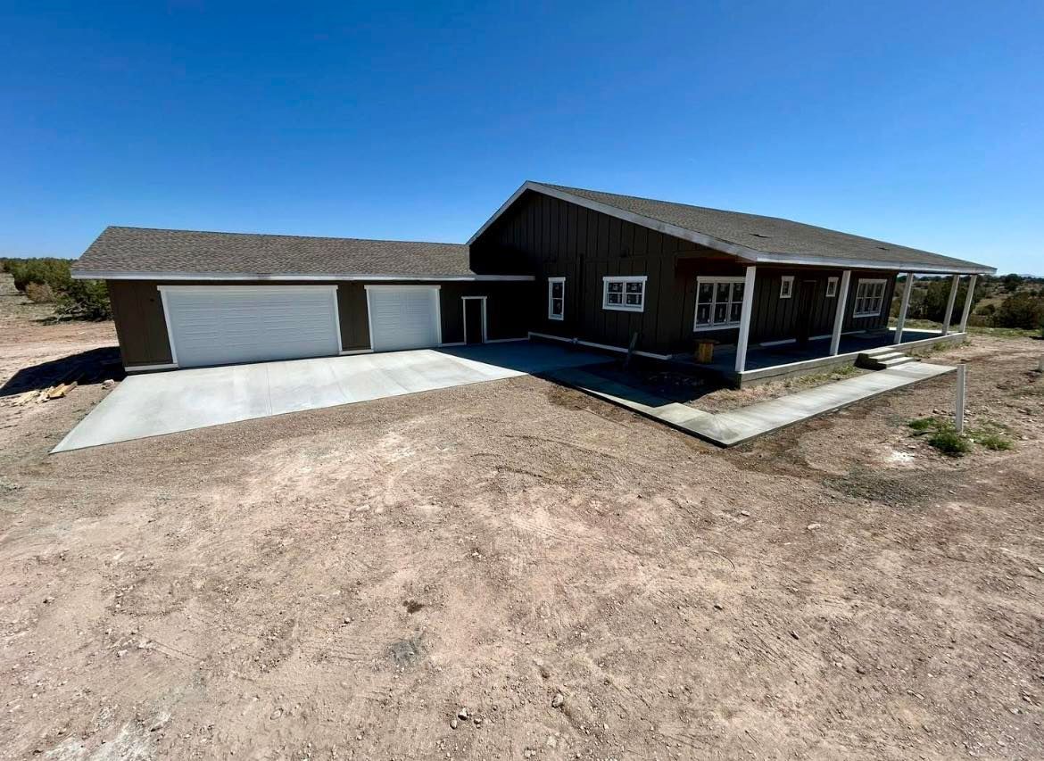 Brown house with two-car garage, concrete driveway, and covered porch on a sunny, dirt lot.