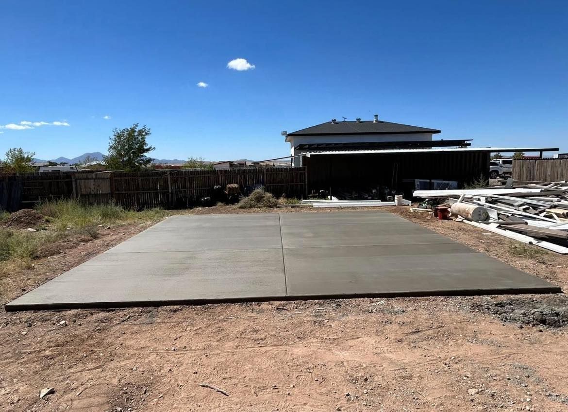 A freshly poured concrete pad in a yard, with a house and blue sky in the background.