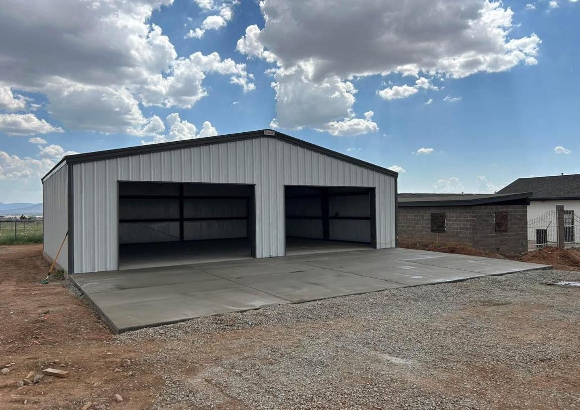 Metal garage with two bays, gray concrete pad, and gravel ground. Blue sky with clouds in the background.