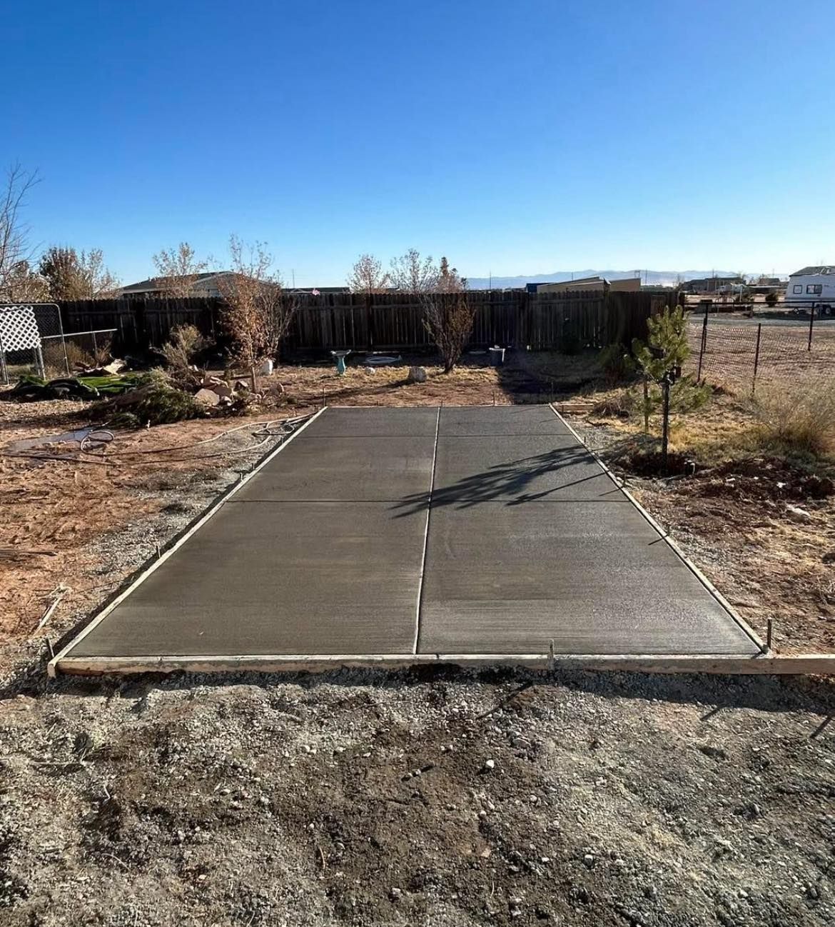 A newly poured, wet concrete slab in a yard, framed by wooden boards. Sunny day, blue sky.