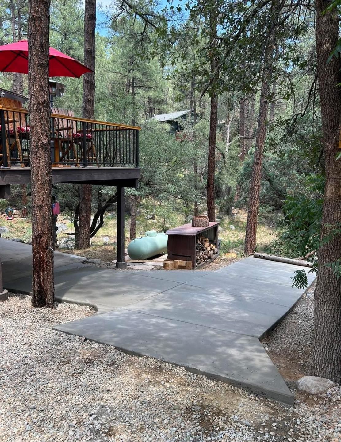Concrete patio with deck, trees. Propane tank and firewood near. Red umbrella on deck.