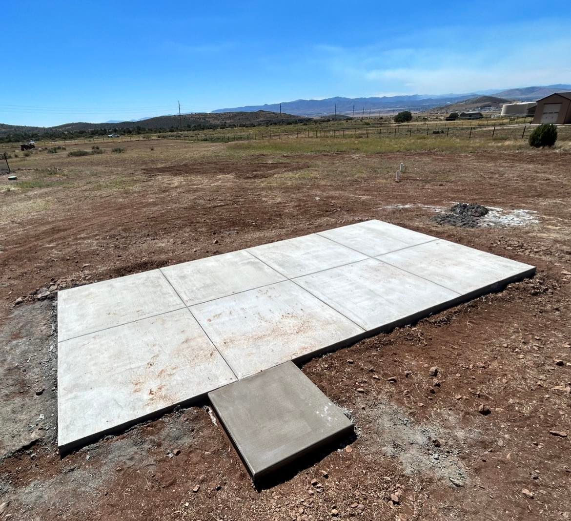 Concrete slabs on dry, dirt ground, ready for construction in an open field, under a blue sky.