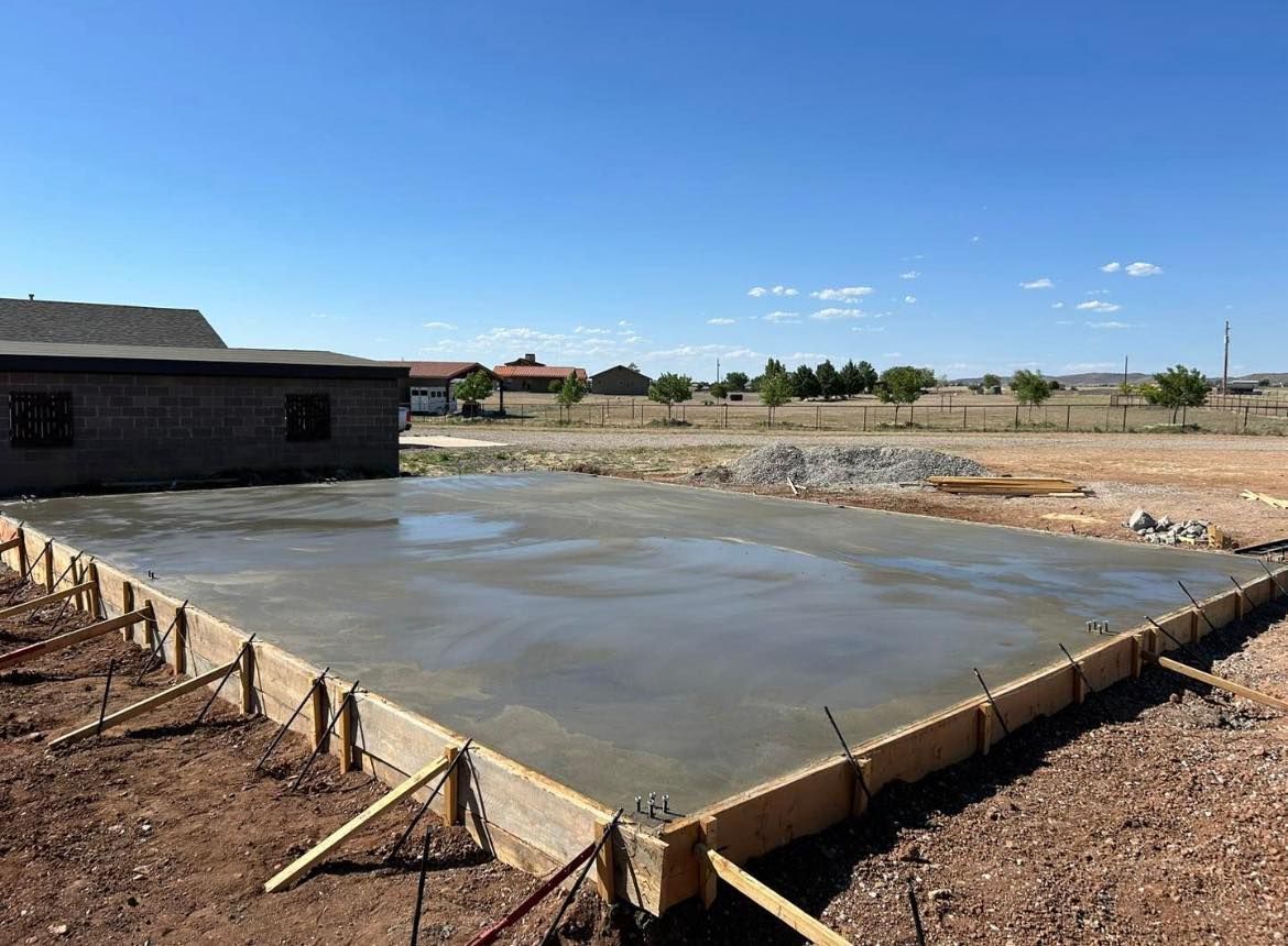 Fresh concrete foundation with wooden framing, outdoors under a blue sky, partially constructed building in the background.