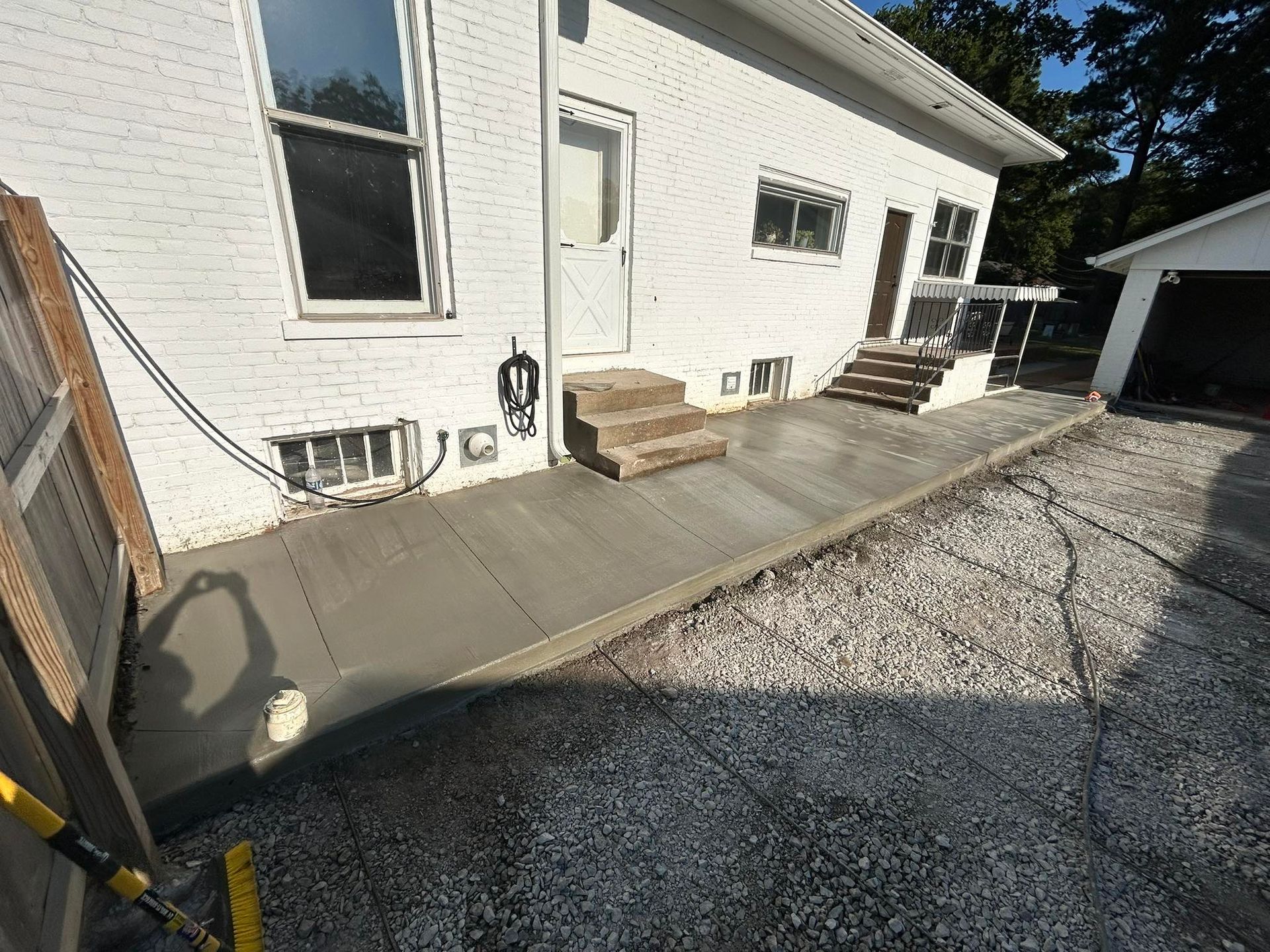 Newly poured concrete patio next to a white house with steps and a door. Gravel driveway.