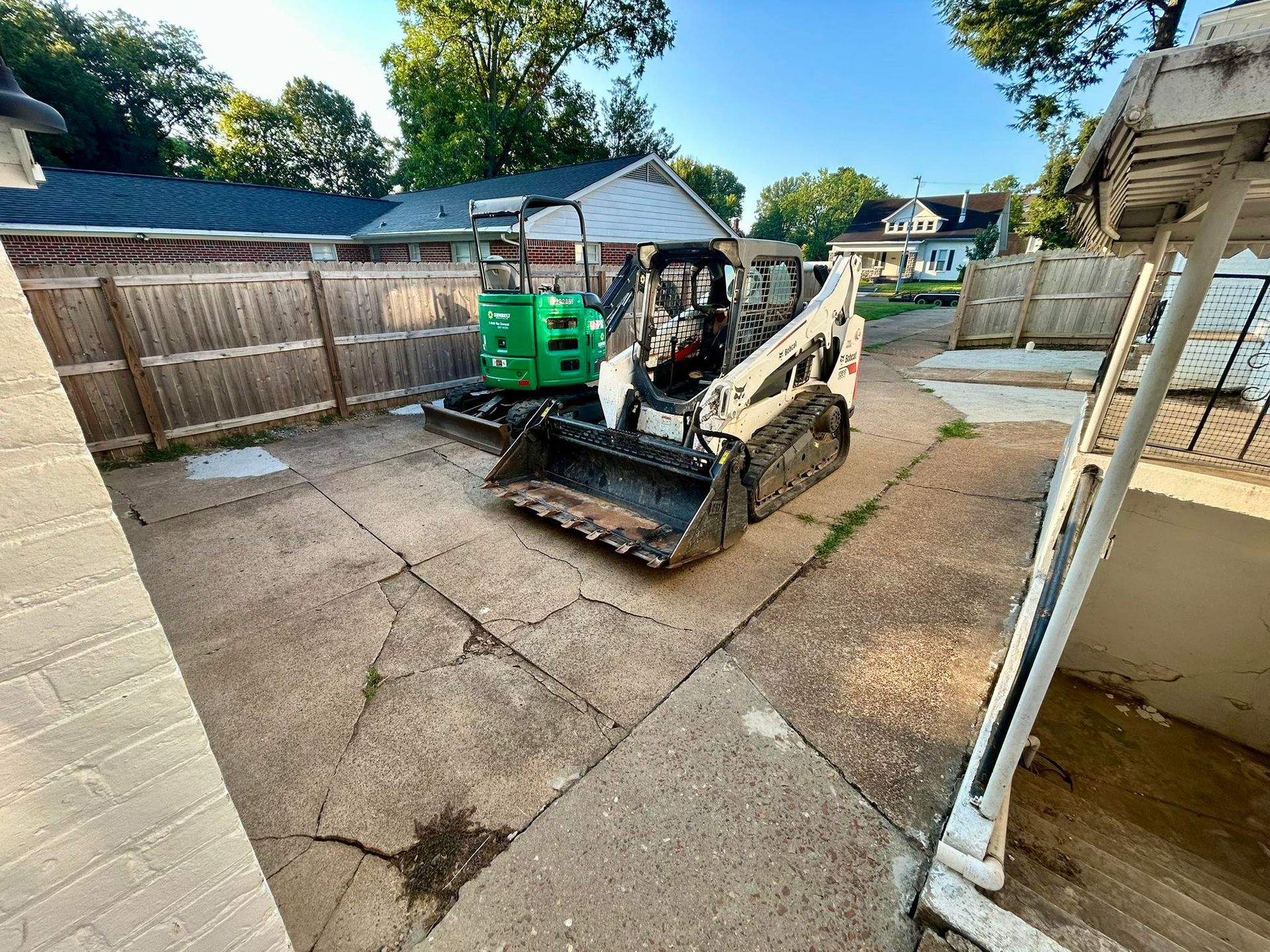 Bobcat skid-steer loader parked on concrete patio near a wooden fence and houses.