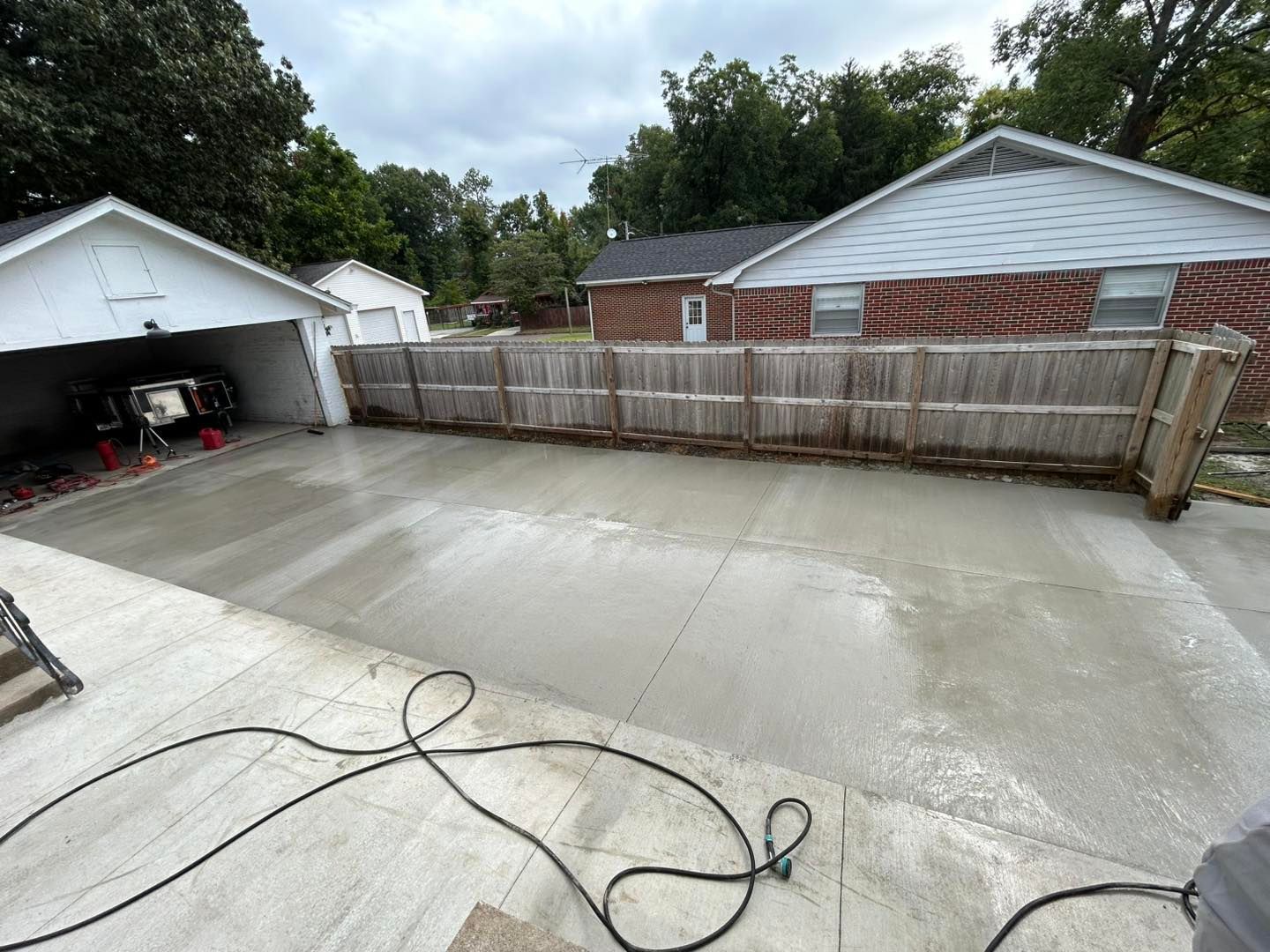 Newly poured concrete driveway with adjacent garage, fence, and houses under a cloudy sky.