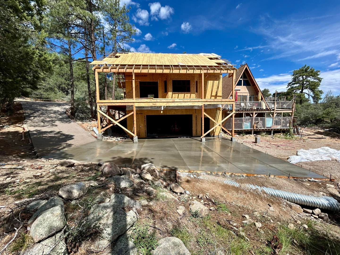 Construction of a two-story wooden house with a garage, wet concrete, and adjacent house on a hillside.