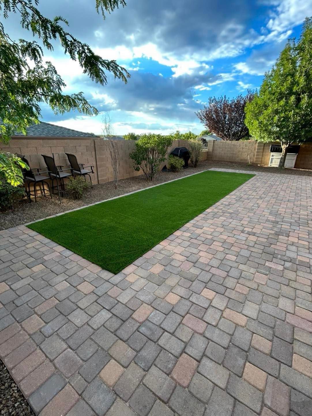 Backyard with artificial grass strip, brick patio, chairs, and cloudy sky.
