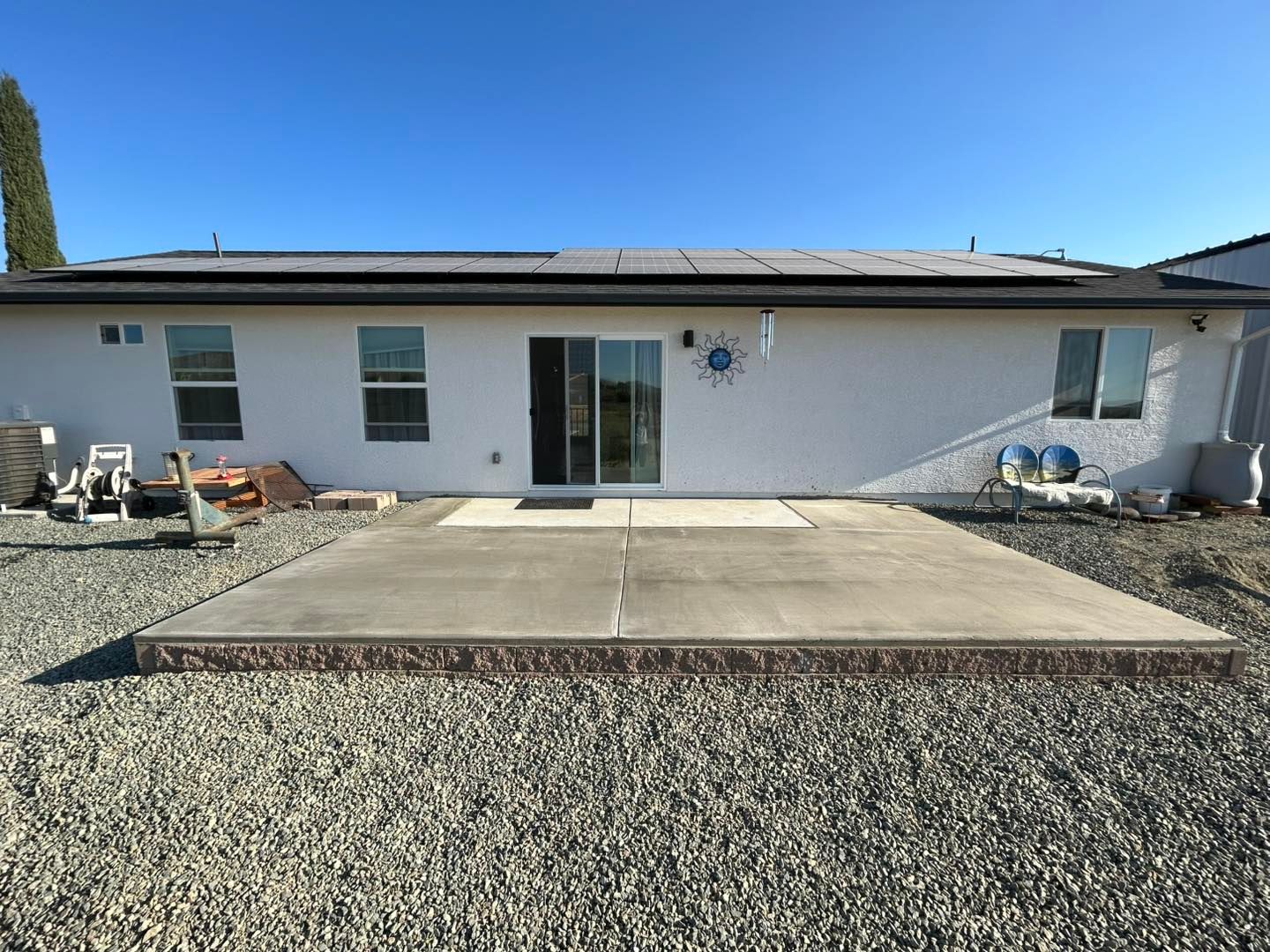 Backyard concrete patio with brown border, in front of a white house with sliding glass door and solar panels.