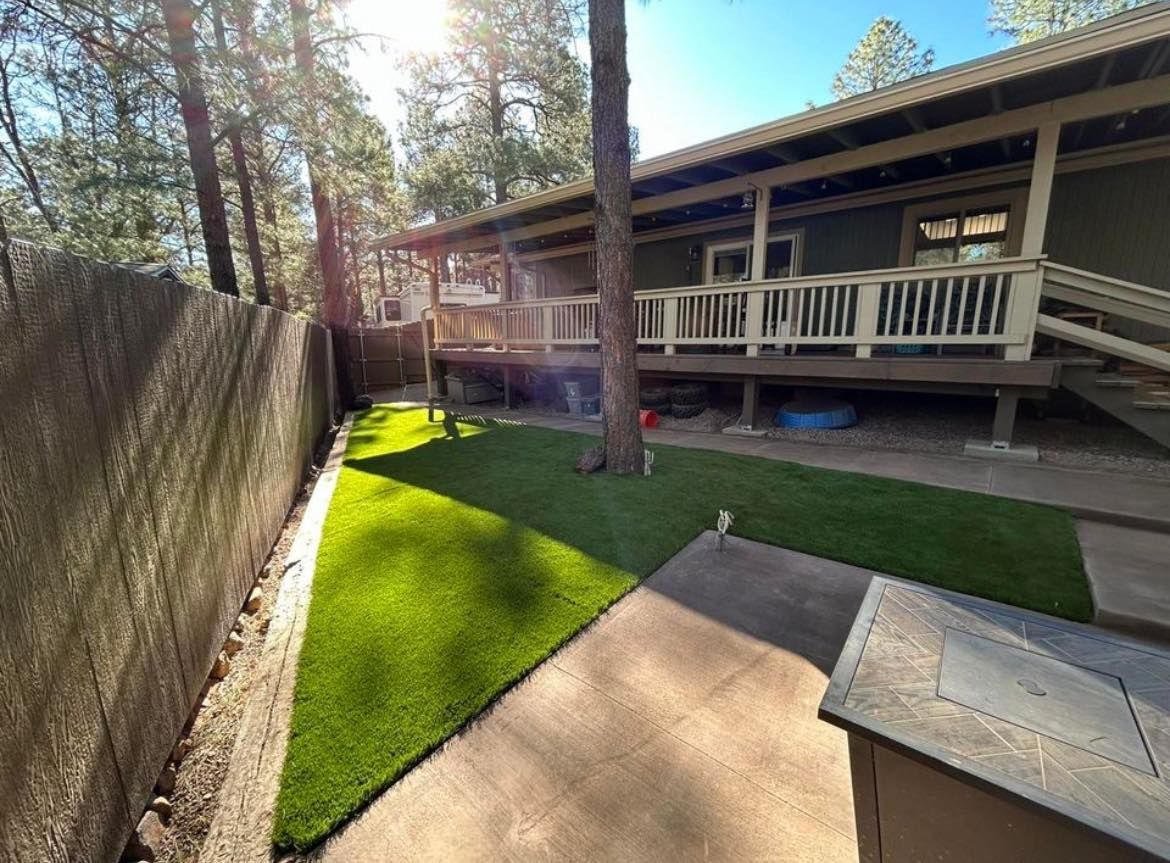 Backyard with green lawn, deck, and trees. Concrete pathway and wooden fence on left. Sunny day.