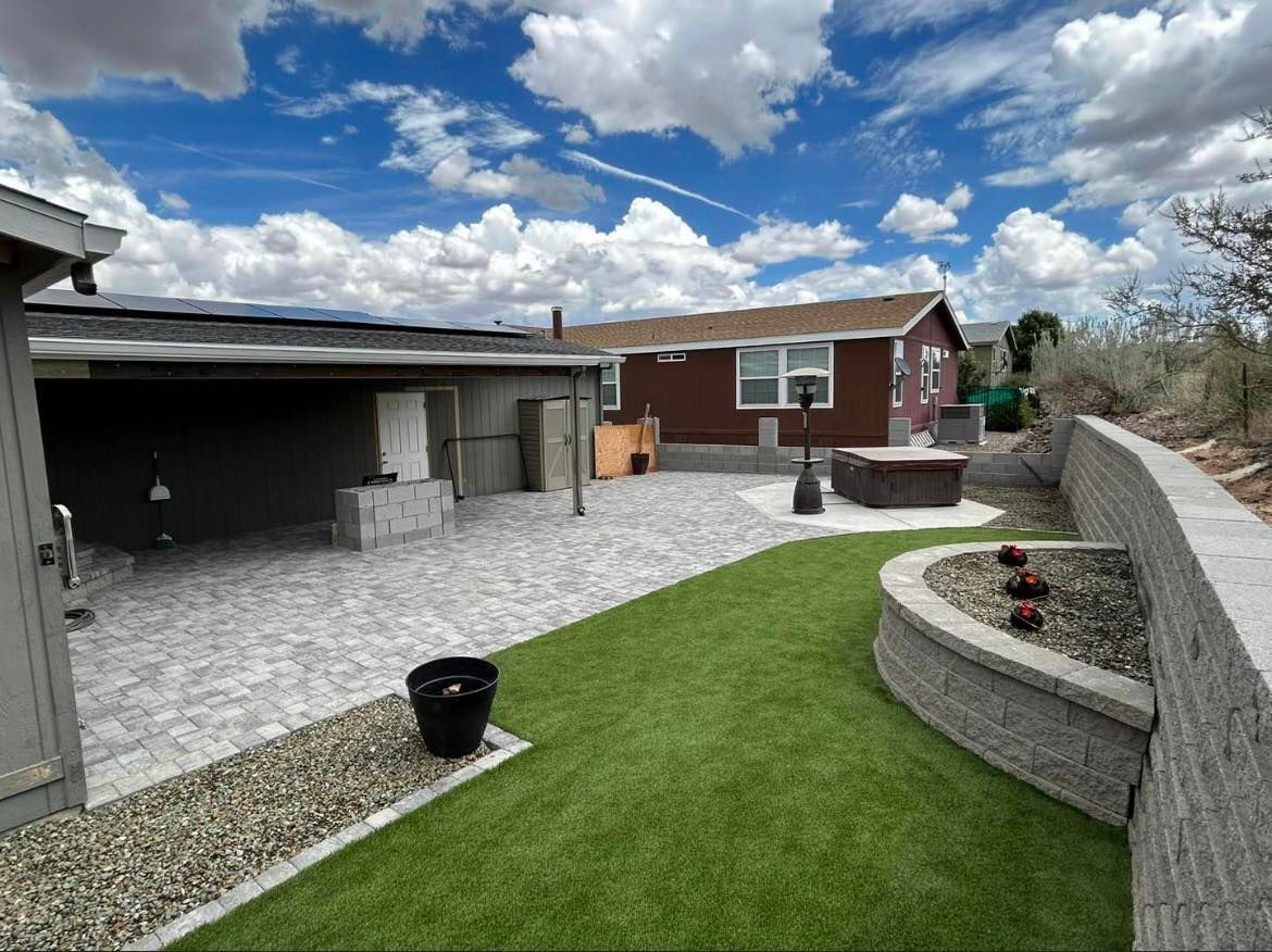 Backyard patio with artificial grass, retaining wall, and hot tub under a blue sky.