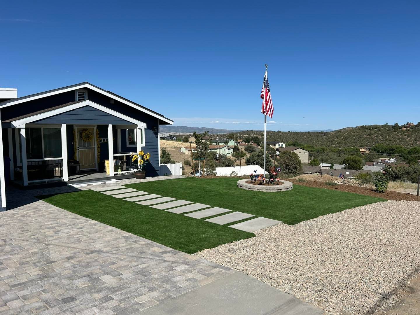 Blue house with American flag, stepping stones, and gravel driveway on a hillside under a blue sky.