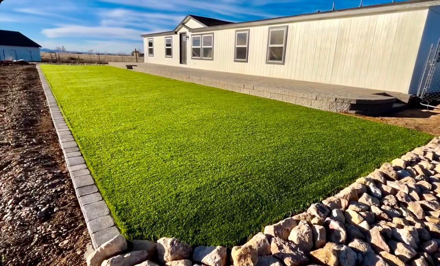 Bright green artificial turf next to a beige house, bordered by stone and gray blocks on a sunny day.