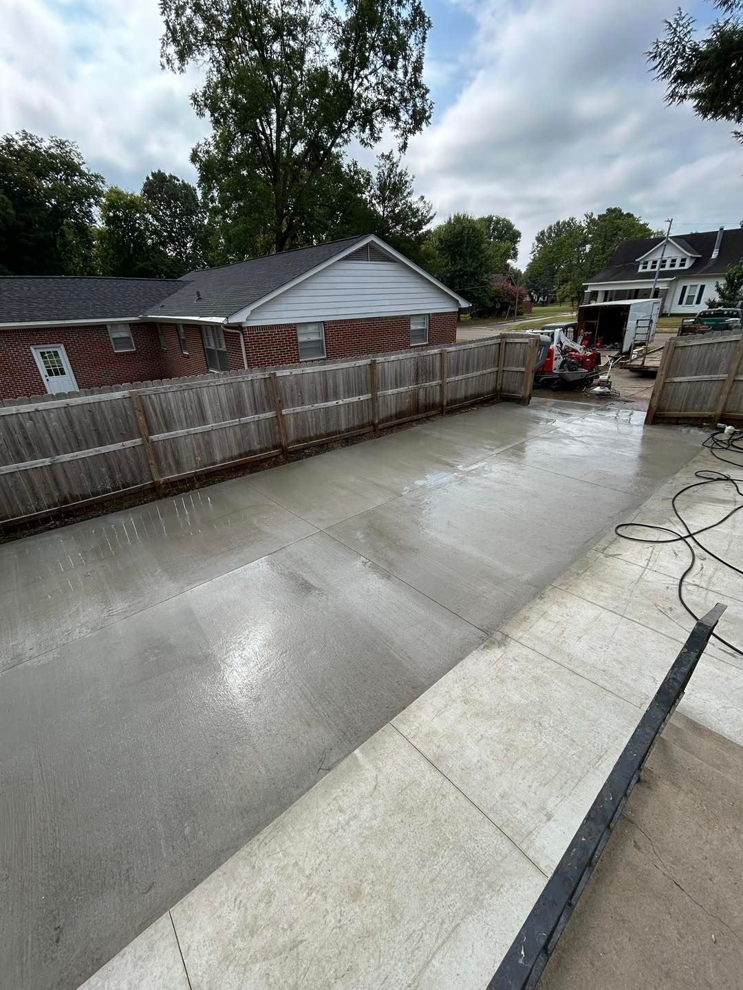 Freshly poured concrete driveway bordered by a wooden fence, with buildings and equipment in the background under a cloudy sky.