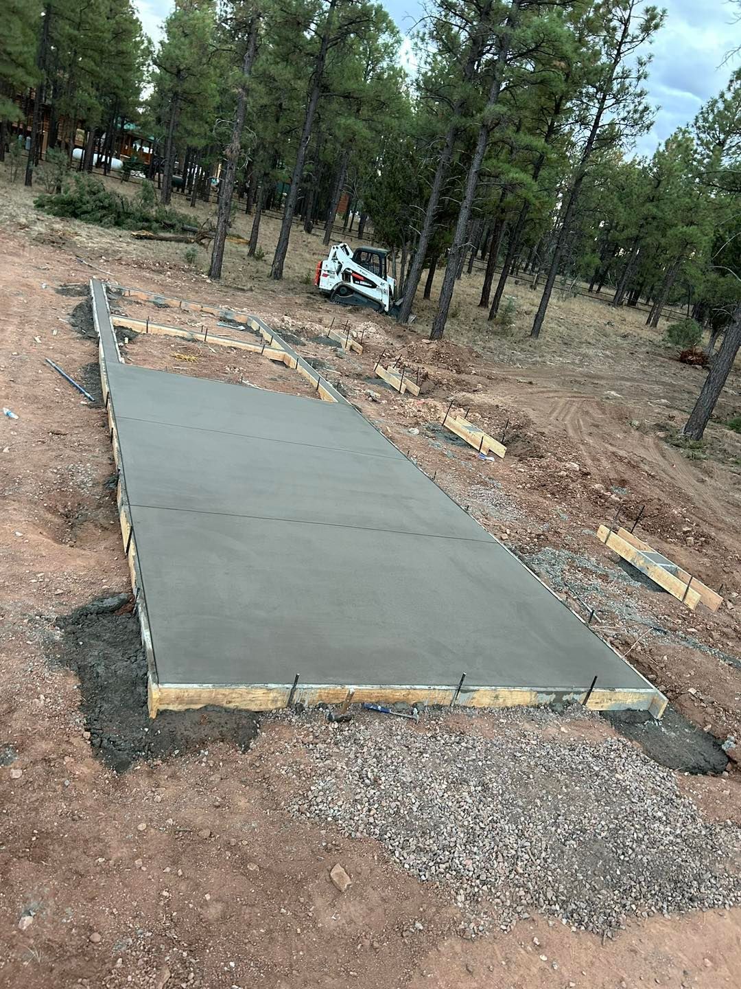 Concrete foundation being poured on a gravel base, bordered by wooden forms, with trees in the background.
