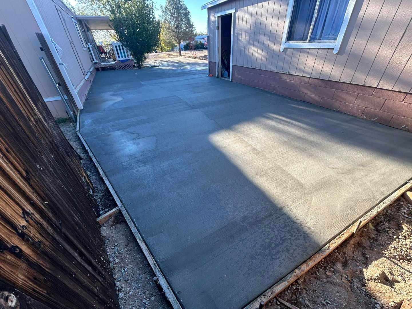 Freshly poured concrete patio next to a weathered wooden fence and building with open shed door.