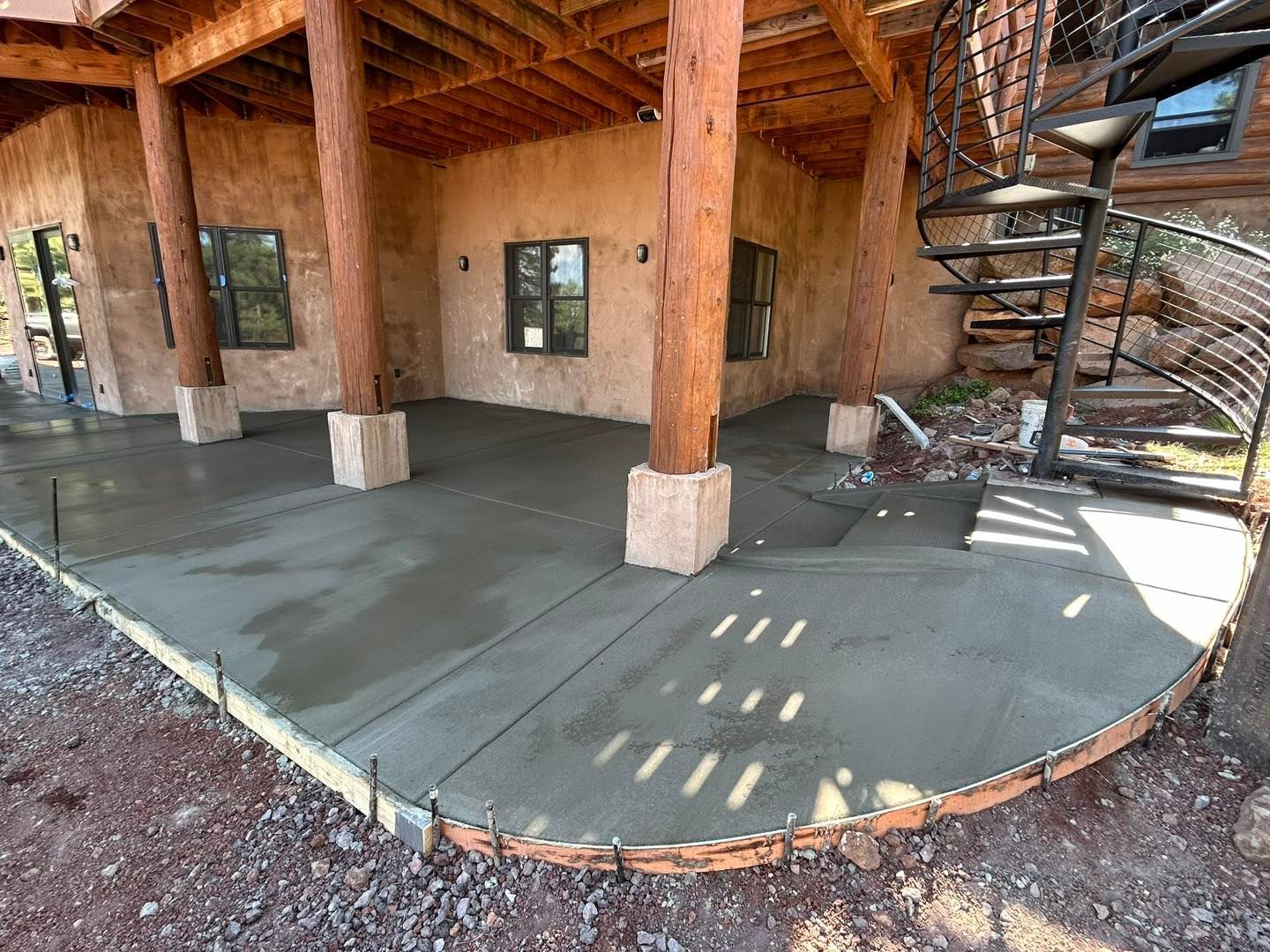 Newly poured concrete patio under a building with wooden support beams and a spiral staircase.