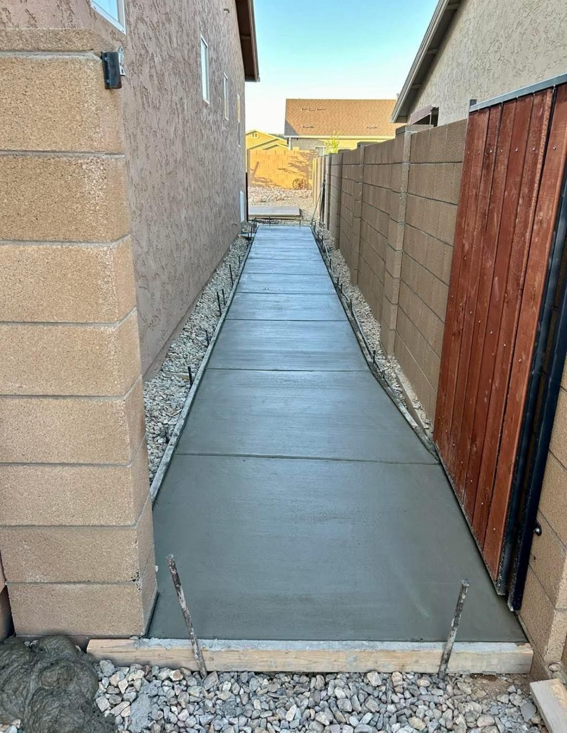 A narrow, newly poured concrete walkway between two walls, with gravel borders and a wooden gate.