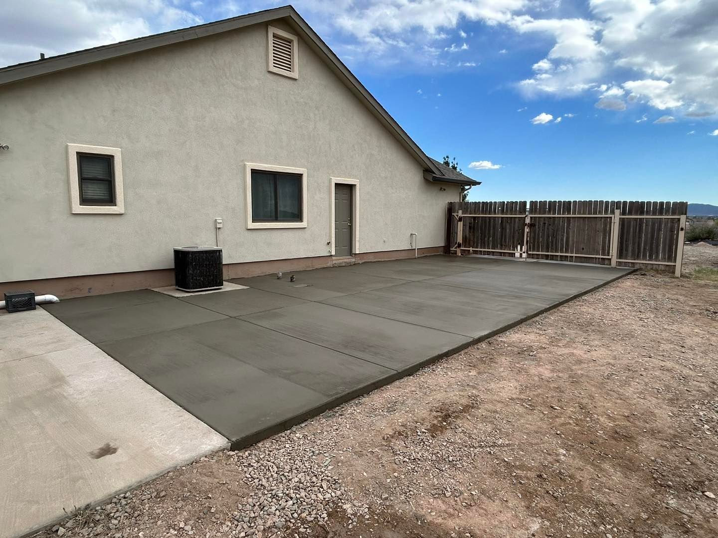 Newly poured concrete patio next to a house with beige stucco walls, set in a dry, desert-like environment.