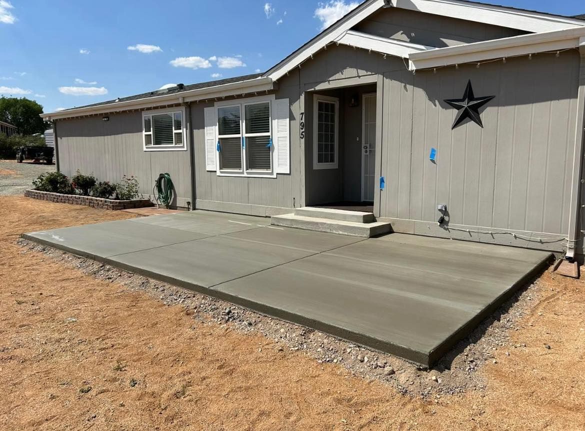 New concrete patio in front of a gray house, surrounded by gravel and bare earth under a blue sky.