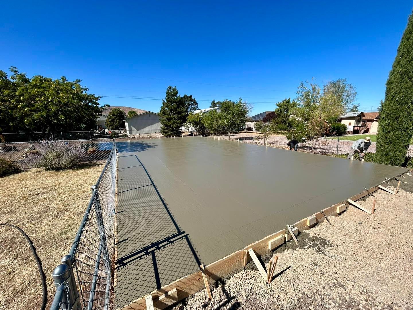 Freshly poured concrete slab bordered by wooden forms and a chain-link fence, outdoors under a clear blue sky.