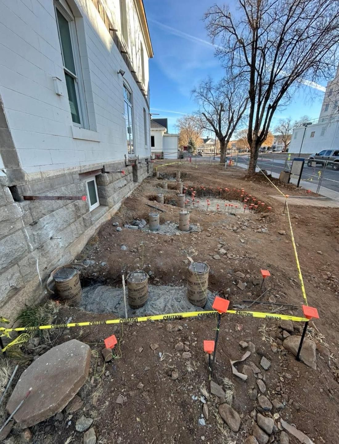 Construction site next to a white building. Footings are being poured. Ground is dirt with construction markers.