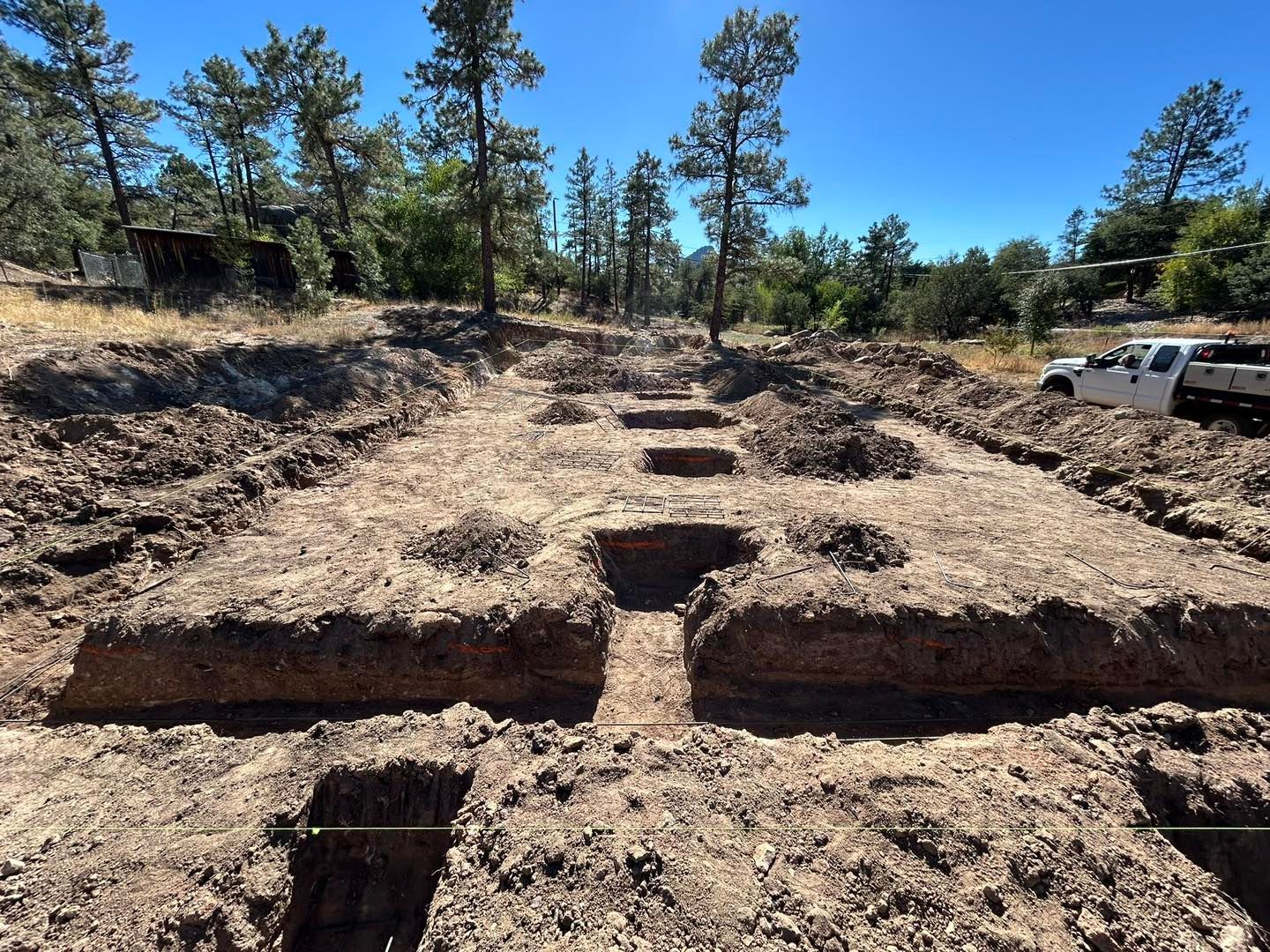 Trenches dug in brown earth for a construction project, under a blue sky with trees in the background.