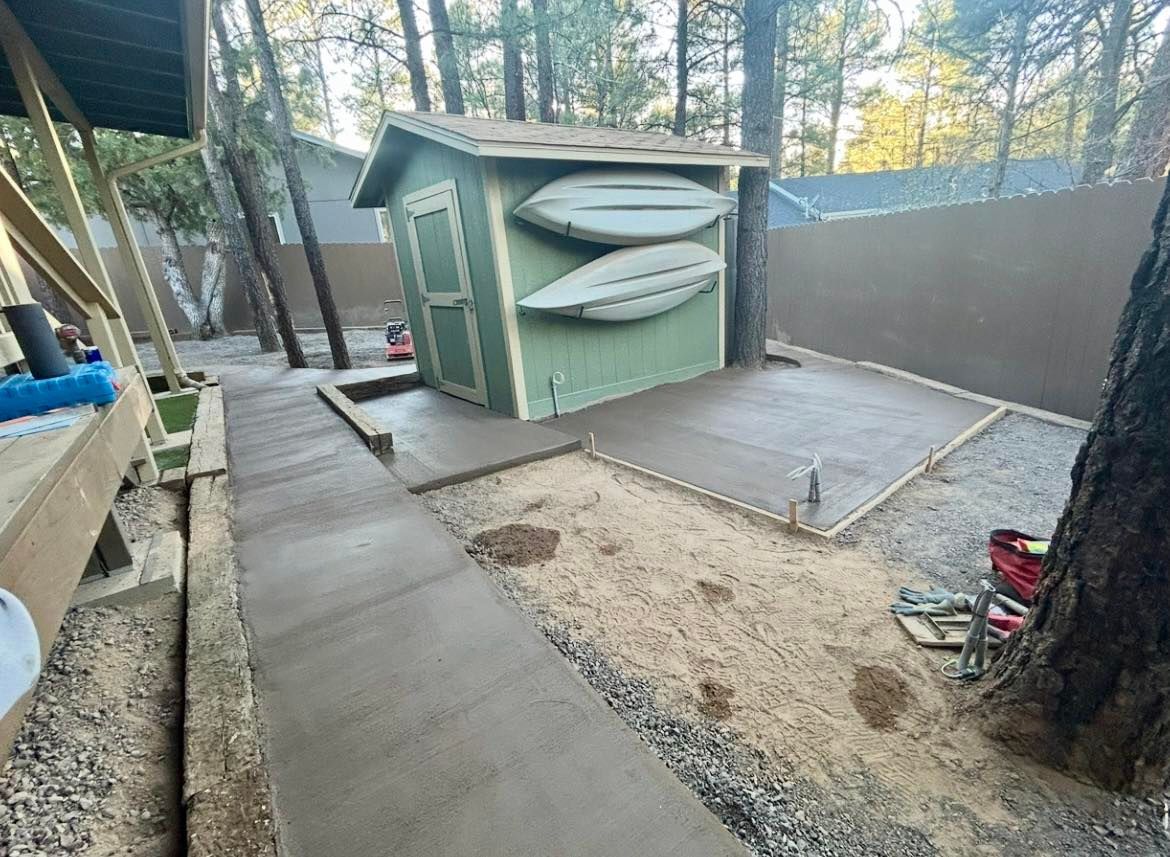 Concrete walkway and patio area with a green shed holding two kayaks, in a wooded setting.