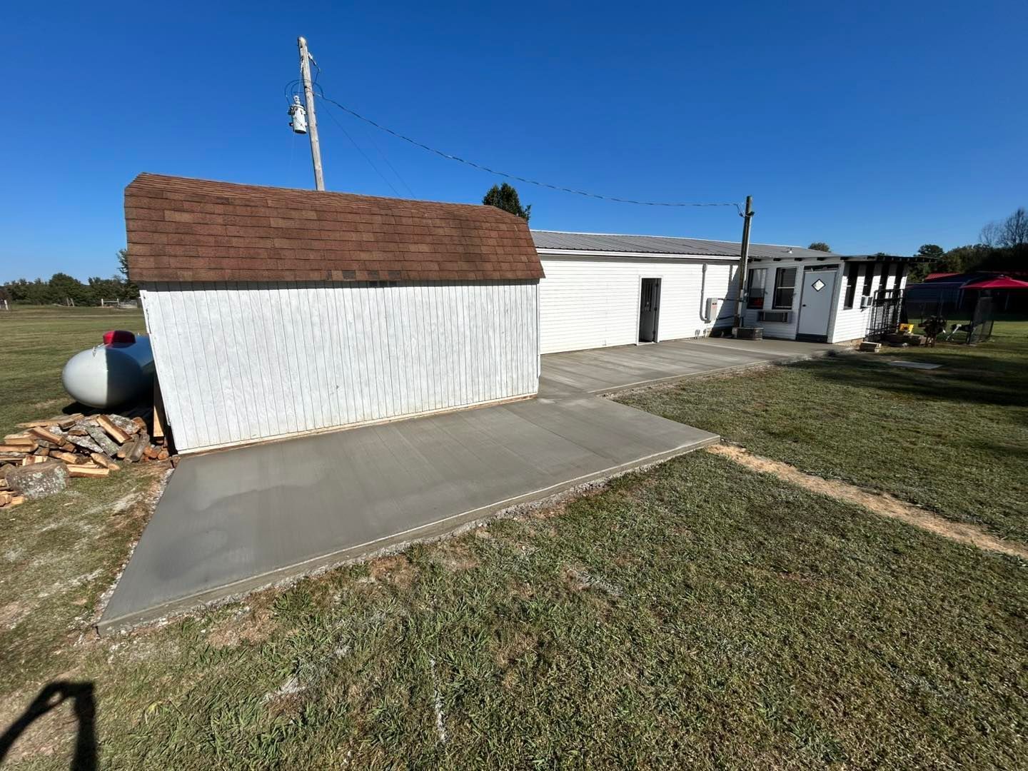 White shed with brown roof next to concrete patio and a white building under a blue sky.