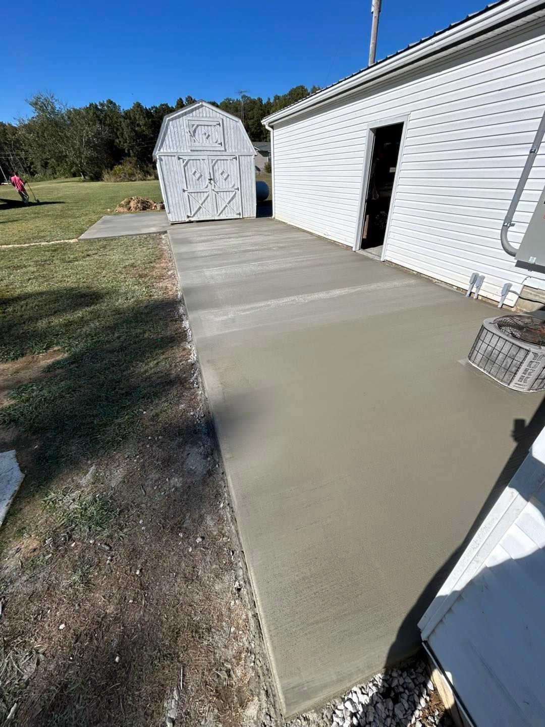 Freshly poured concrete patio next to a white building and a small shed on a sunny day.