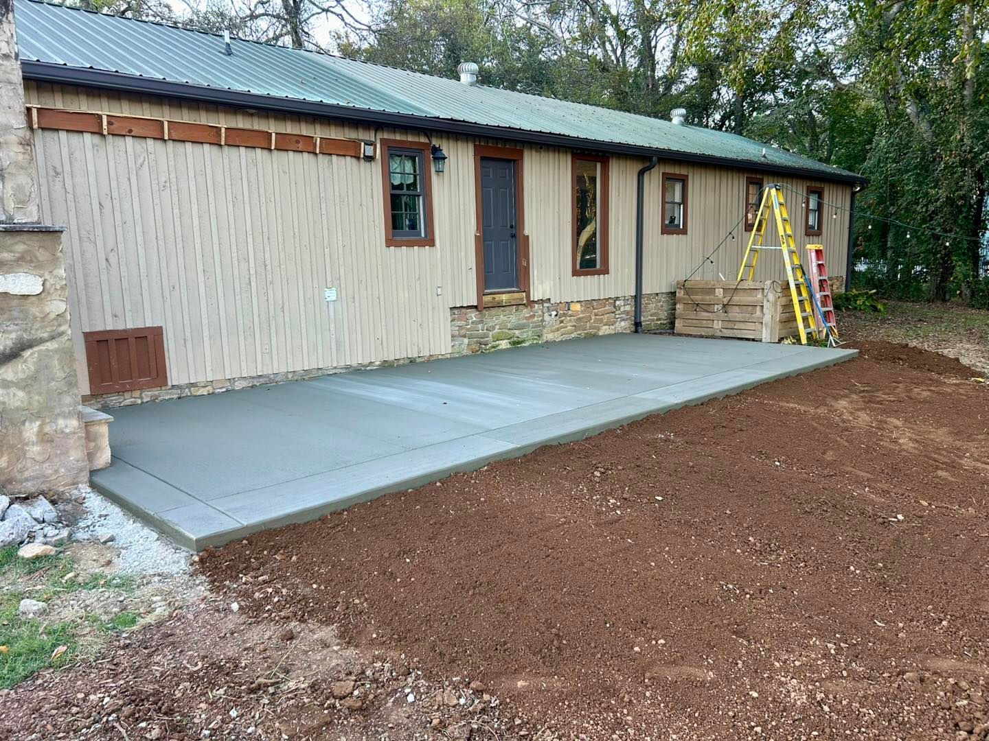 Concrete patio next to a tan house with a green roof, dirt in the foreground.