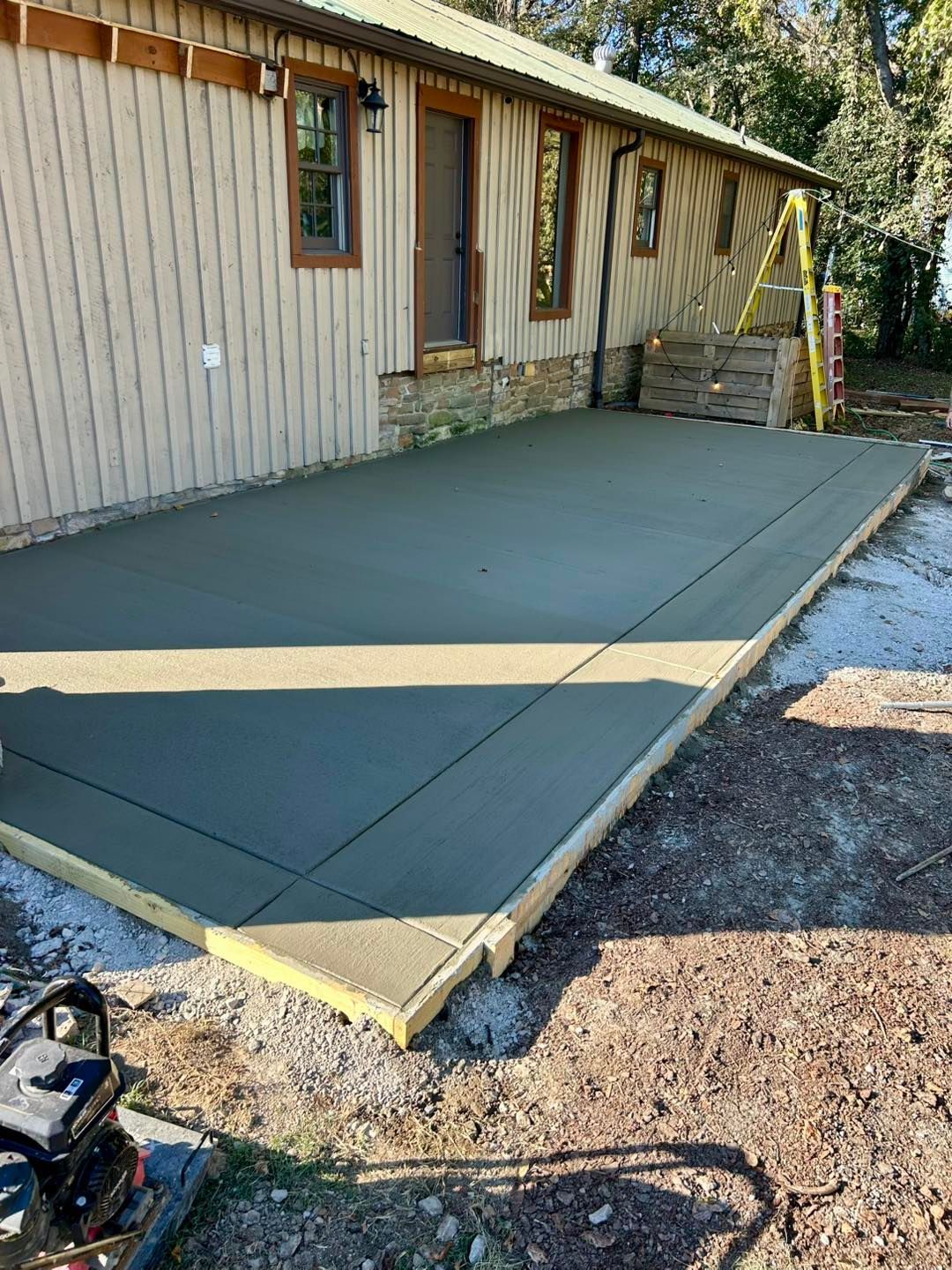 Newly poured concrete patio next to a light brown building. Wooden forms surround the gray slab on gravel.