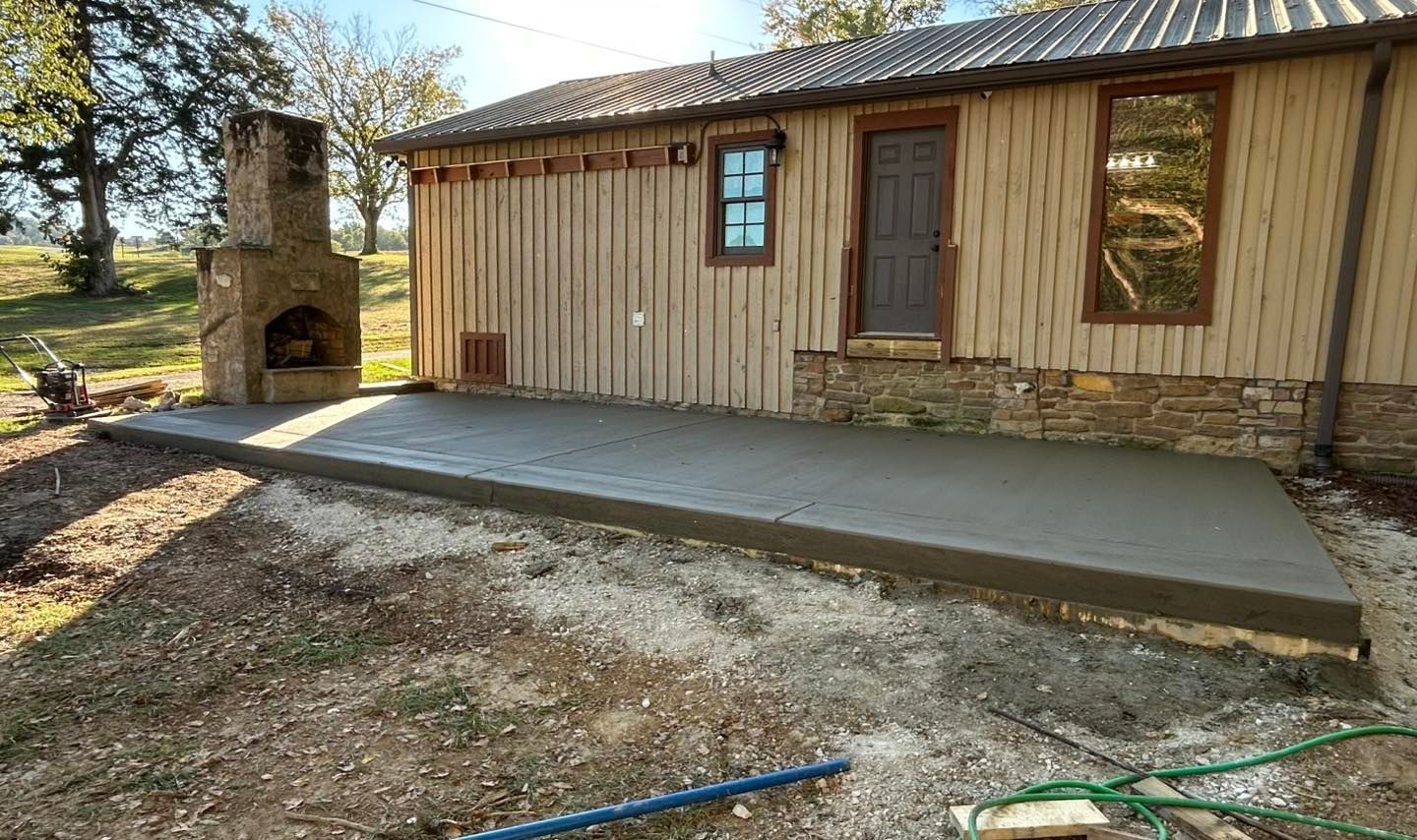 Newly poured concrete patio next to a wooden building with a stone foundation and chimney.