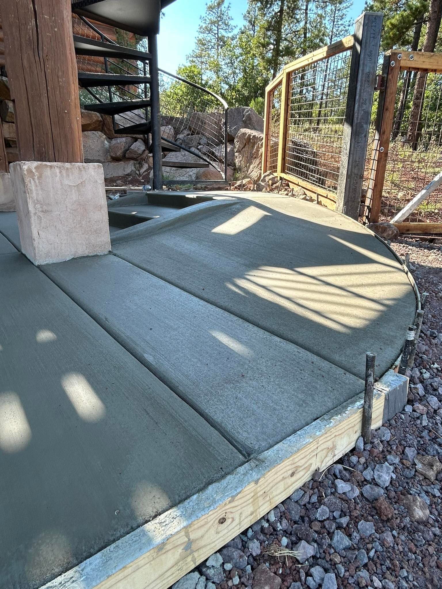 Newly poured concrete patio, bordered by wood, with a spiral staircase and light-colored block in the background.