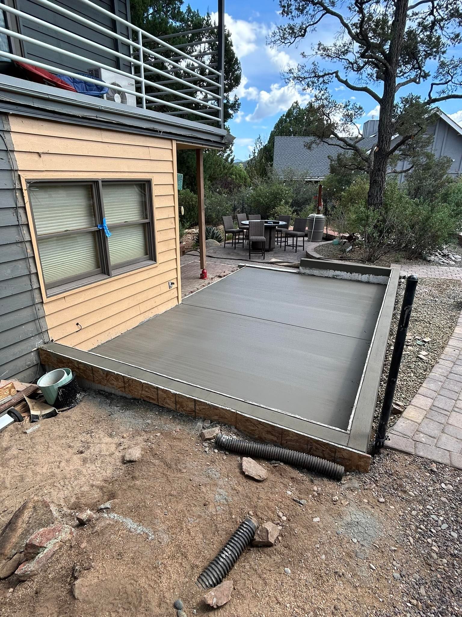 Newly poured concrete patio next to a house with a balcony, set in a yard with trees and outdoor furniture.