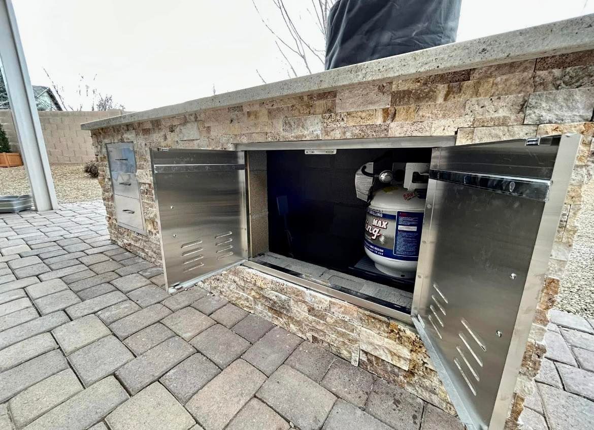 Outdoor stone grill station with stainless steel doors open to reveal a propane tank.
