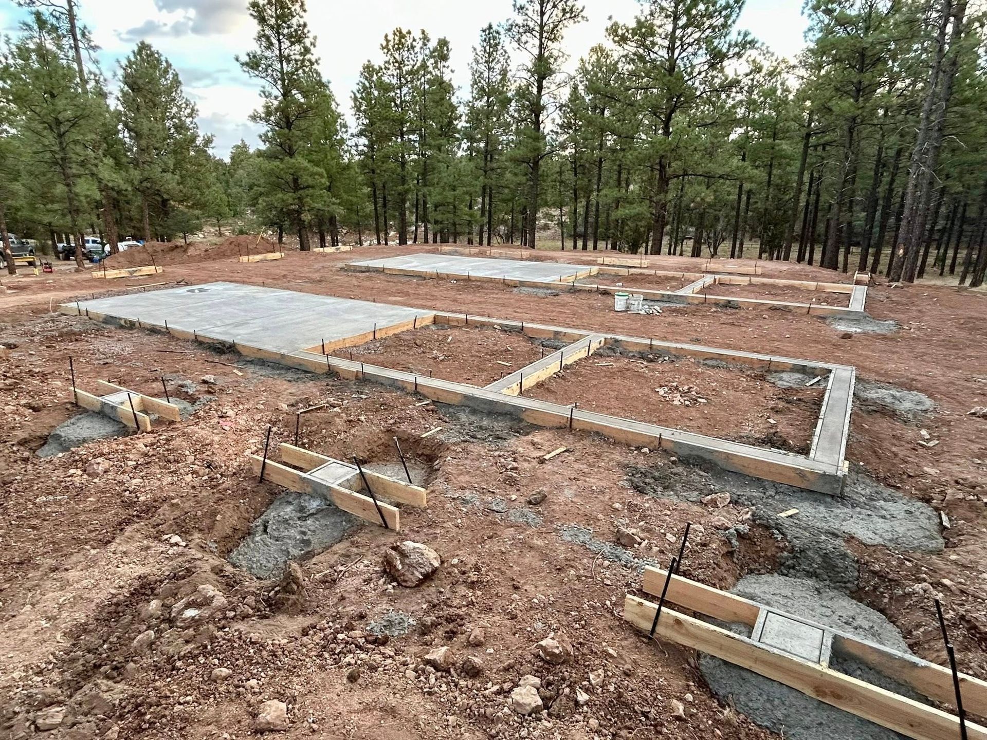 Construction site with concrete foundations and wooden forms, in a wooded area.