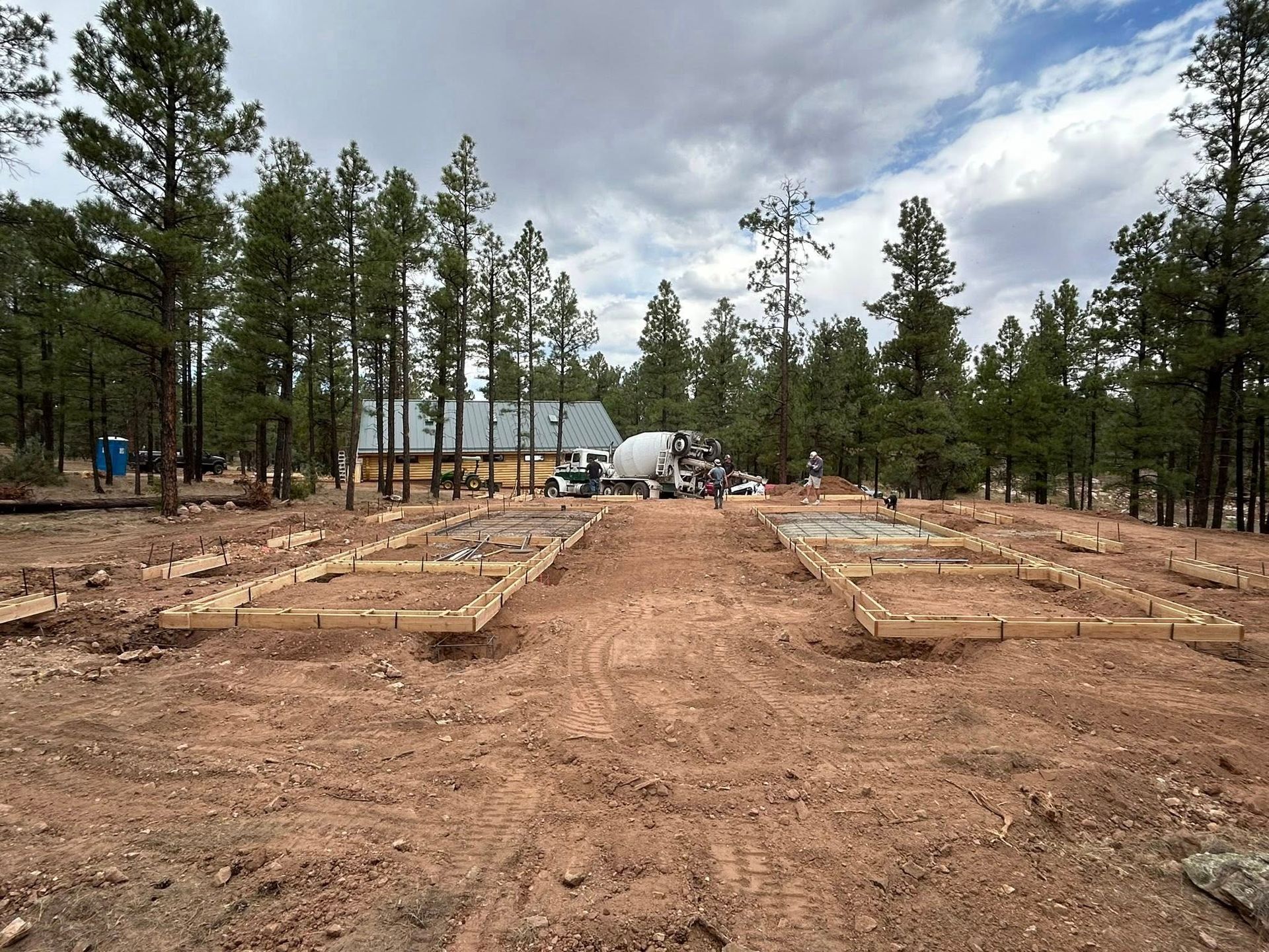 Construction site in a forest with foundation forms, concrete mixer, and building in progress. Cloudy sky.