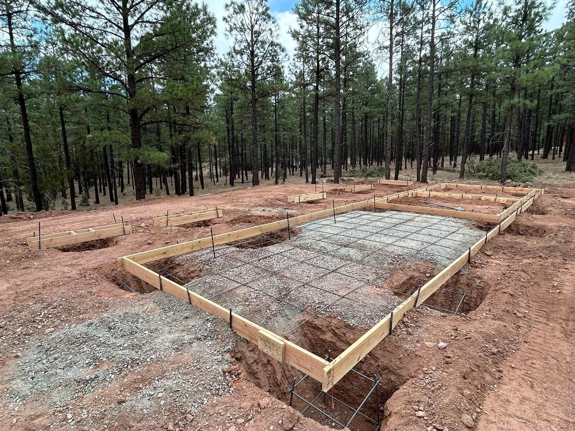 A rectangular construction site with wooden frame, rebar grid, gravel base, and trenches in a forest setting.