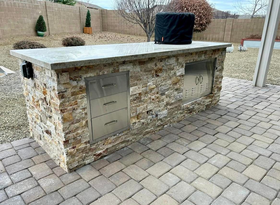 Outdoor kitchen island with stone facade and concrete countertop. Includes grill and storage. Paved patio setting.