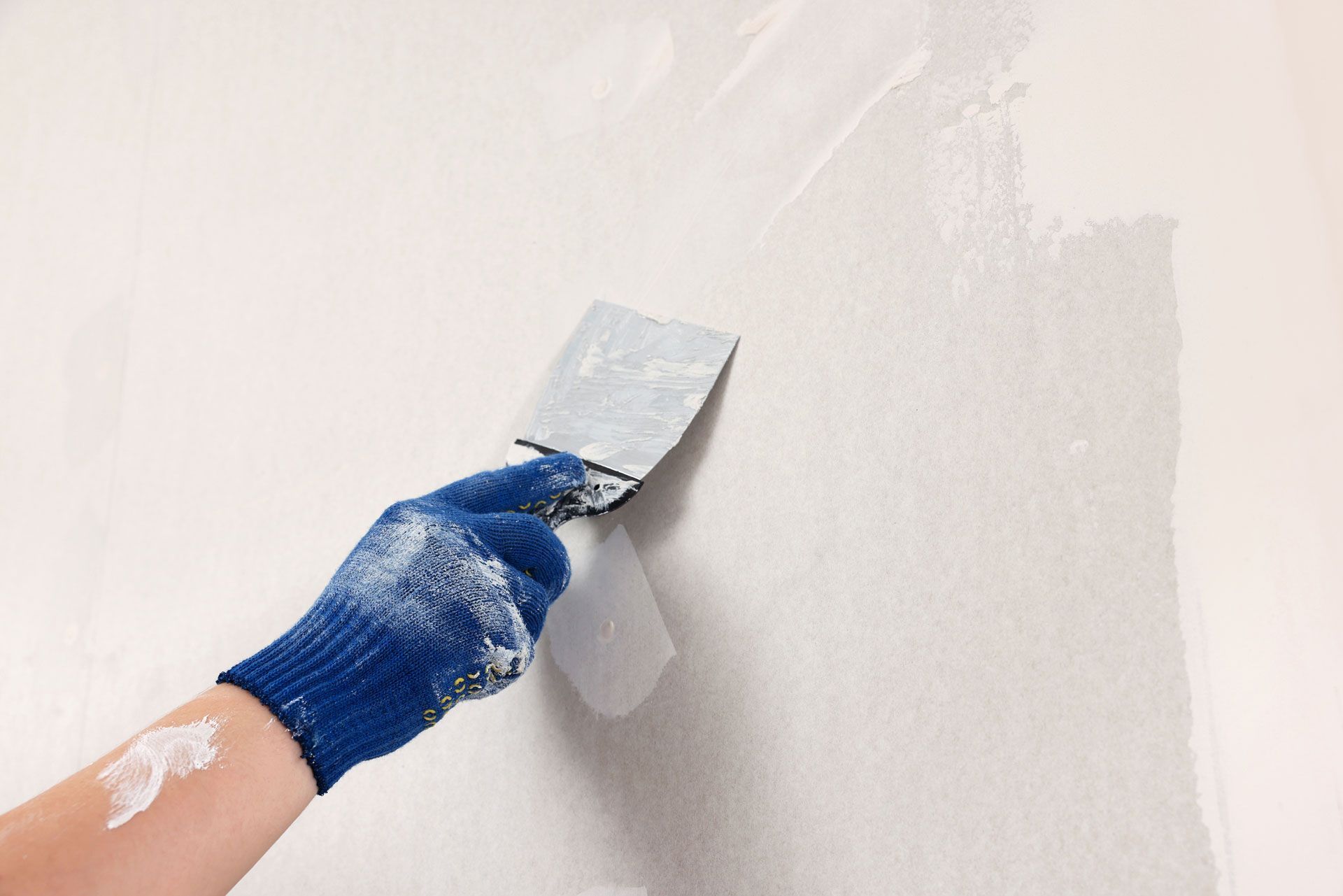 Gloved hand smoothing white paint on a wall with a putty knife
