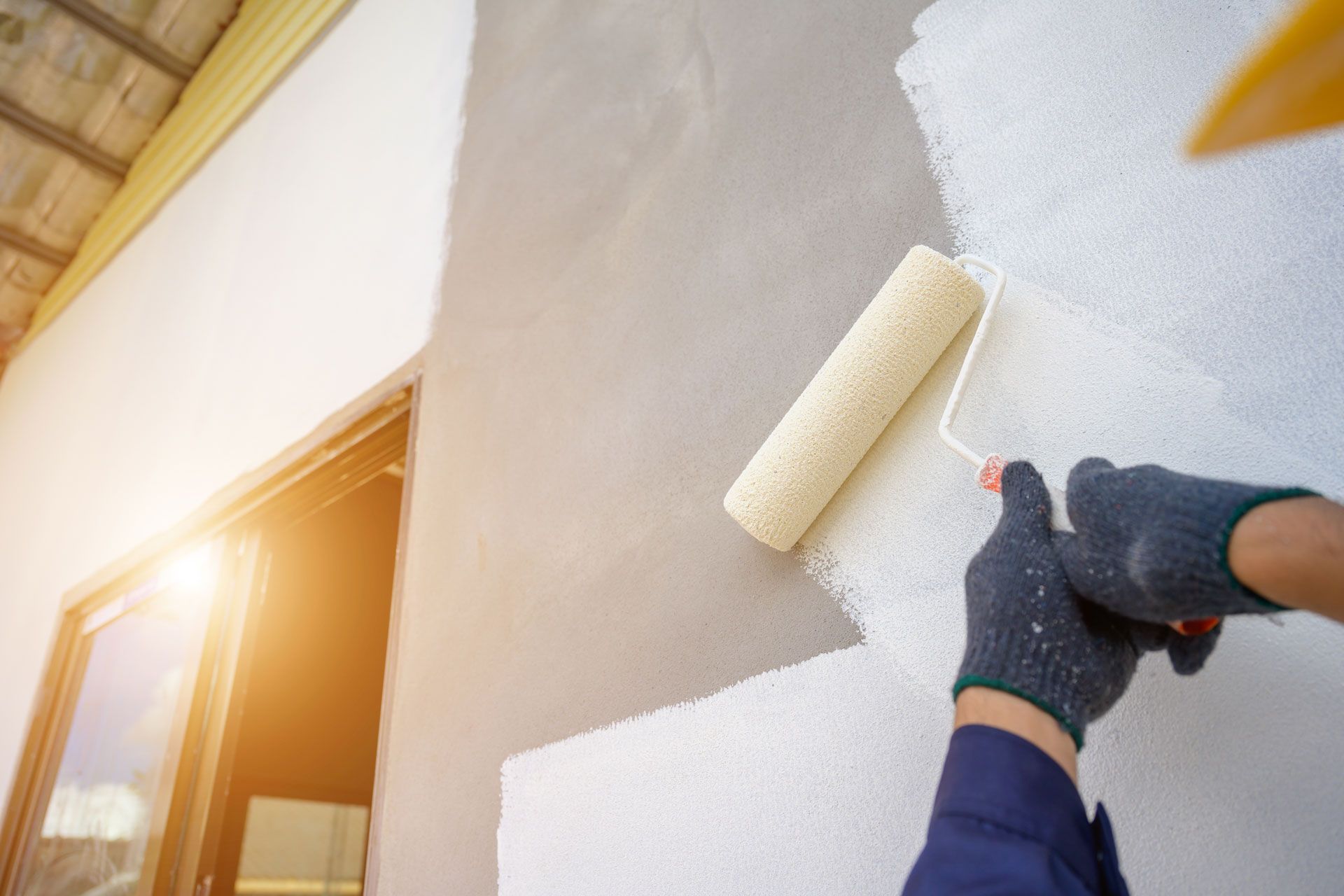 Person painting a white exterior wall with a roller beside a sunlit window
