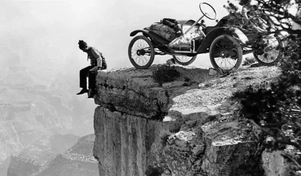 A man is sitting on the edge of a cliff next to an old car.