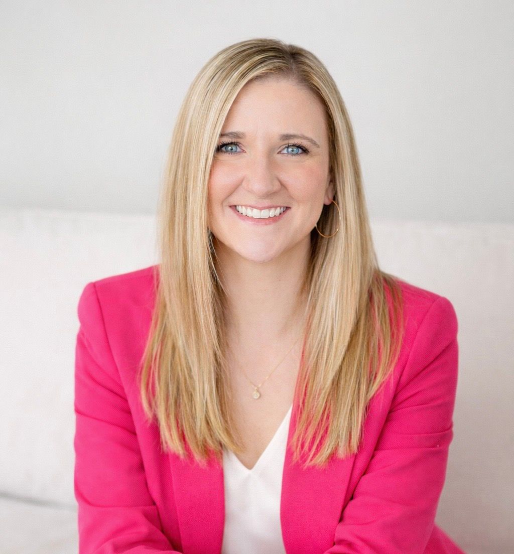 Blonde woman smiling, wearing a pink blazer over a white top; sitting indoors.