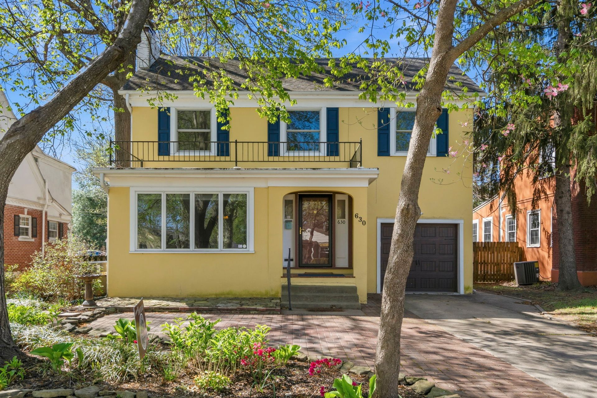 Yellow colonial style home with pink flower and hosta landscaping and green trees with blue skies