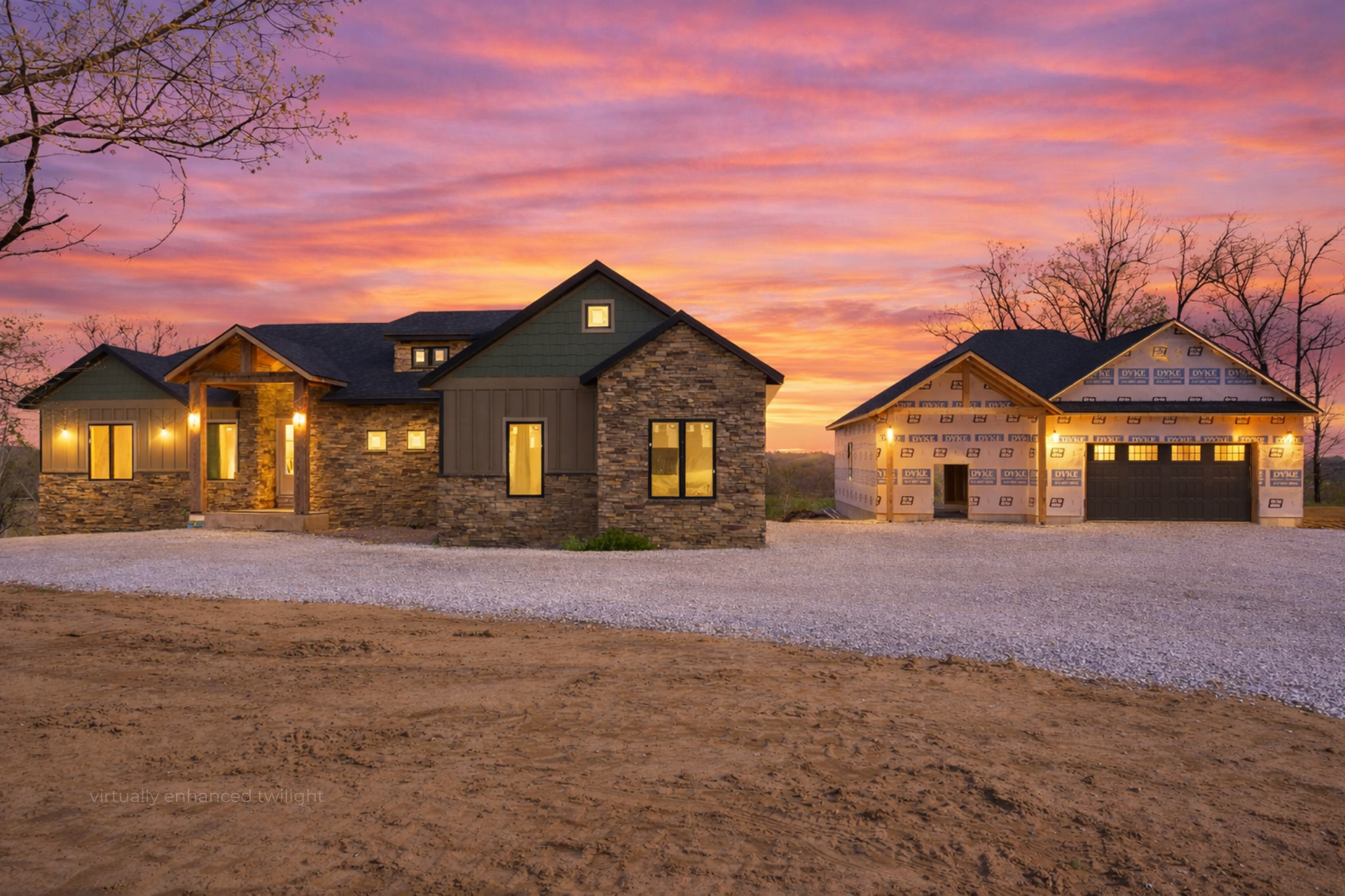 Stone house and detached garage at sunset, with warm lights and a gravel driveway under a pink-orange sky
