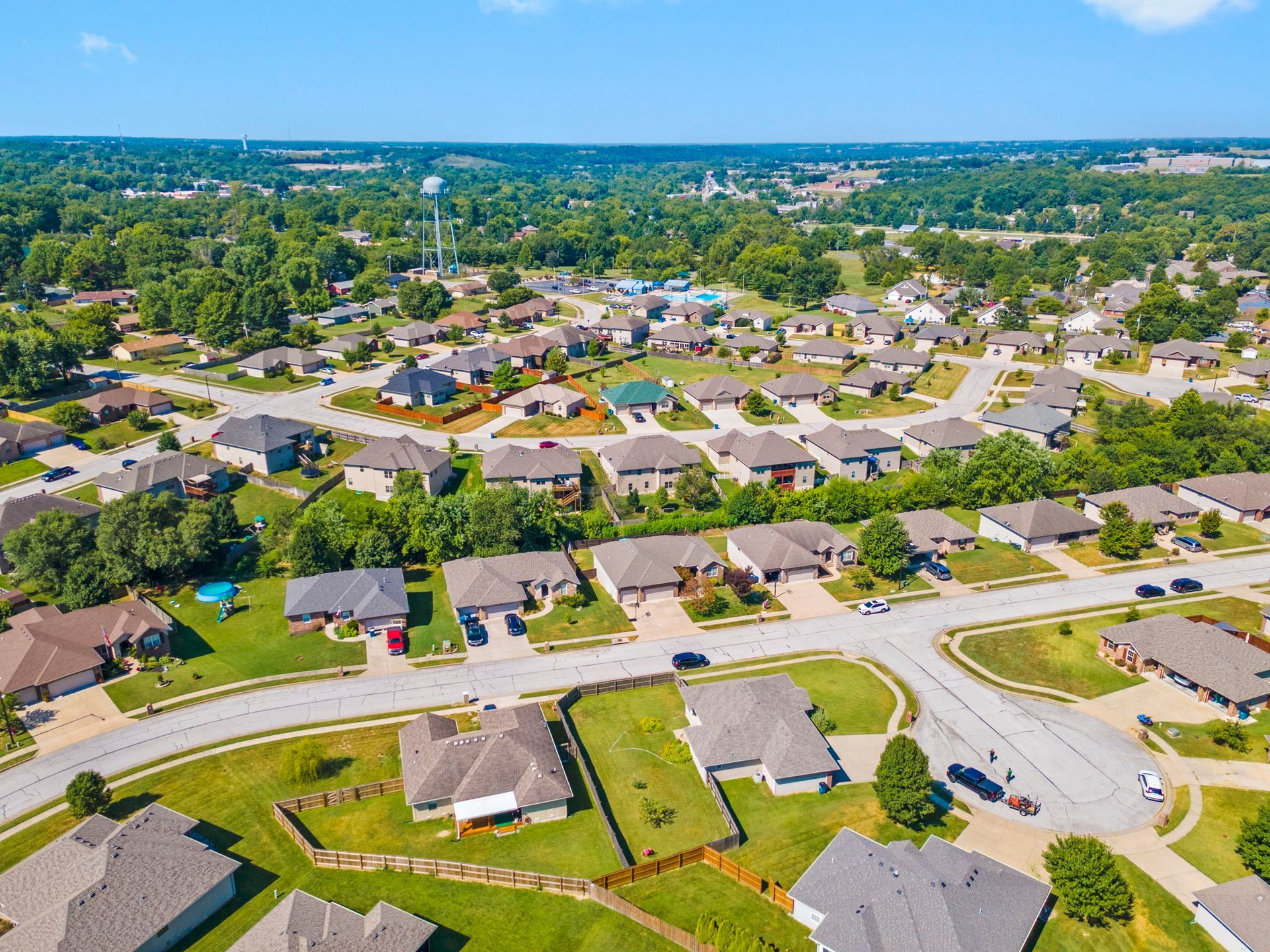 Aerial view of a suburban neighborhood with houses, streets, trees, and a water tower under a blue sky.