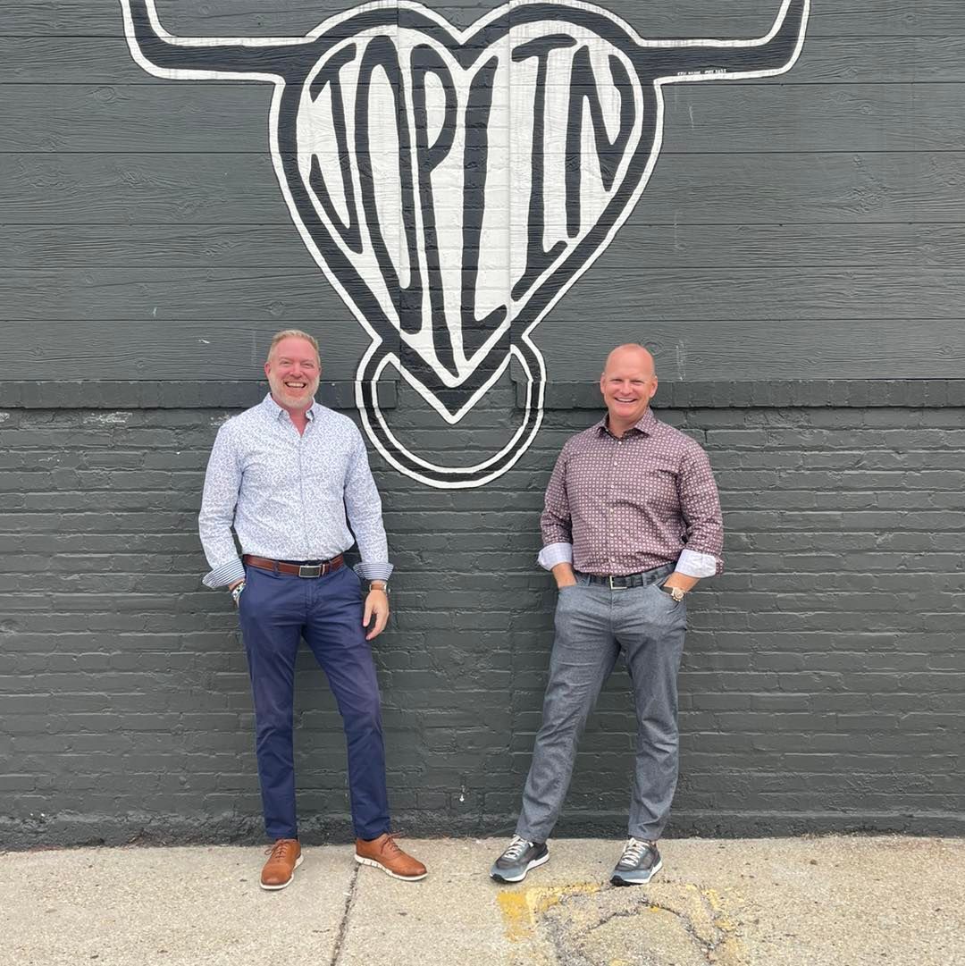 Man with sunglasses gives a thumbs up, carrying a child on his shoulders, both smiling in a park.
