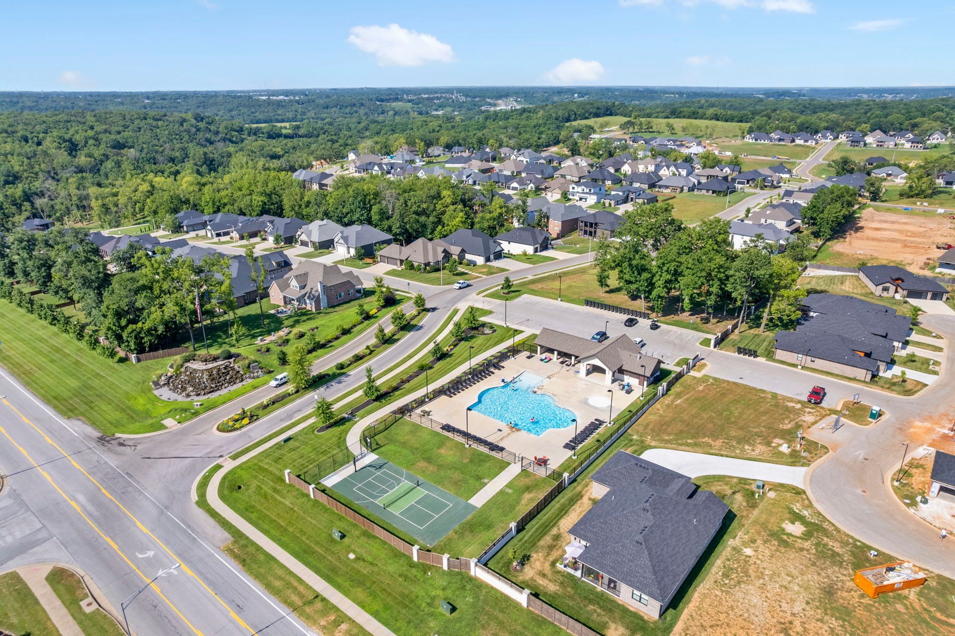 Aerial view of a suburban neighborhood with houses, a pool, tennis courts, and a road on a sunny day.